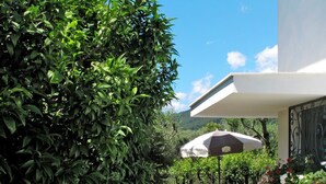 Et moderne udendørsområde med velplejet græsplæne, to stribede liggestole, en stor parasol samt forskellige planter og blomster.