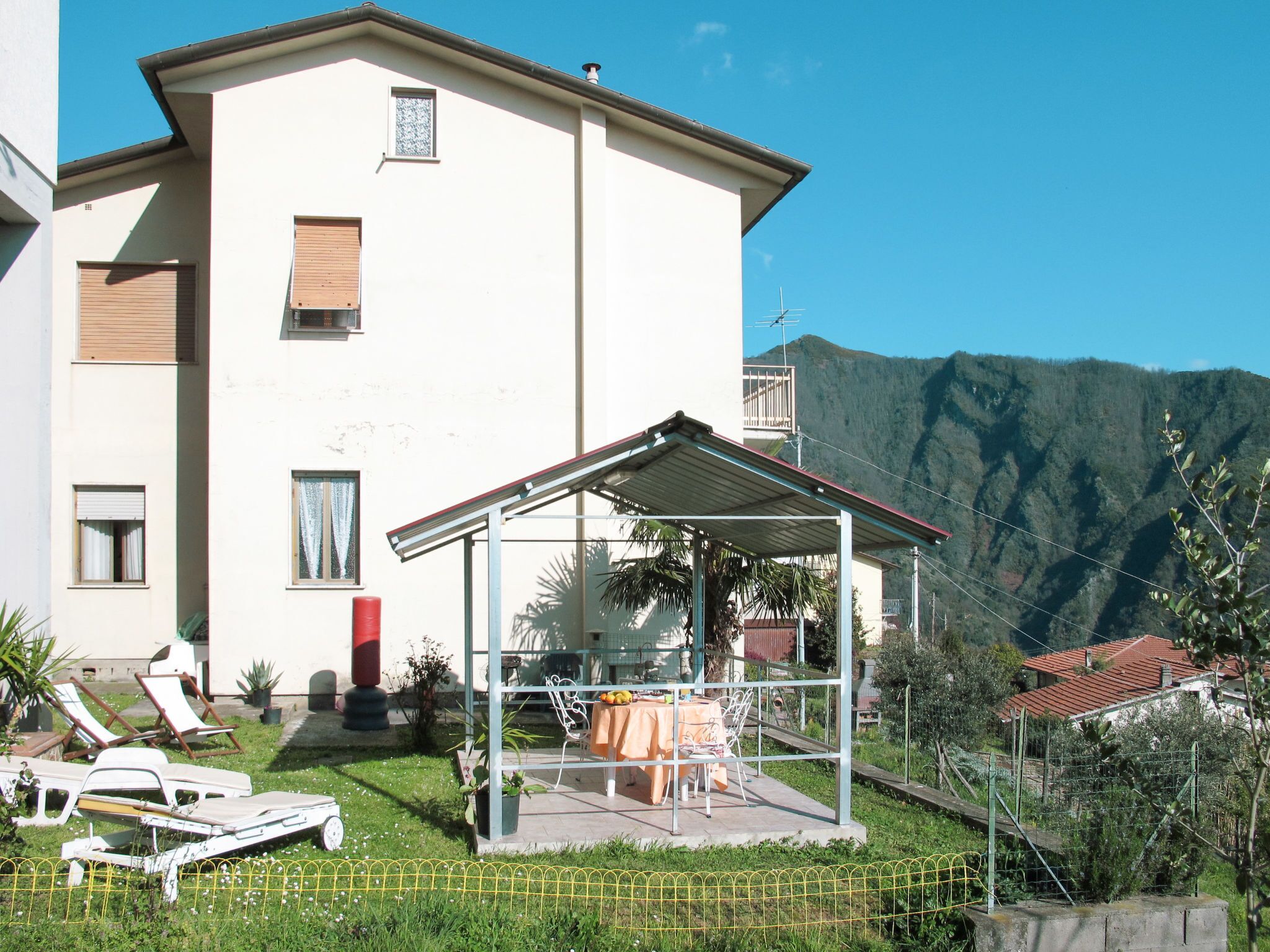 Un edificio de dos pisos con un porche cubierto, un telón de fondo montañoso y un jardín cercado.