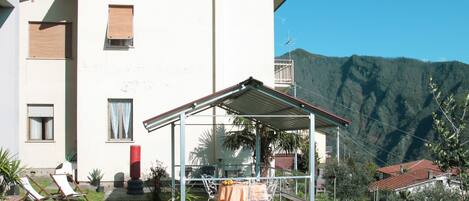Un edificio de dos pisos con un porche cubierto, un telón de fondo montañoso y un jardín cercado.