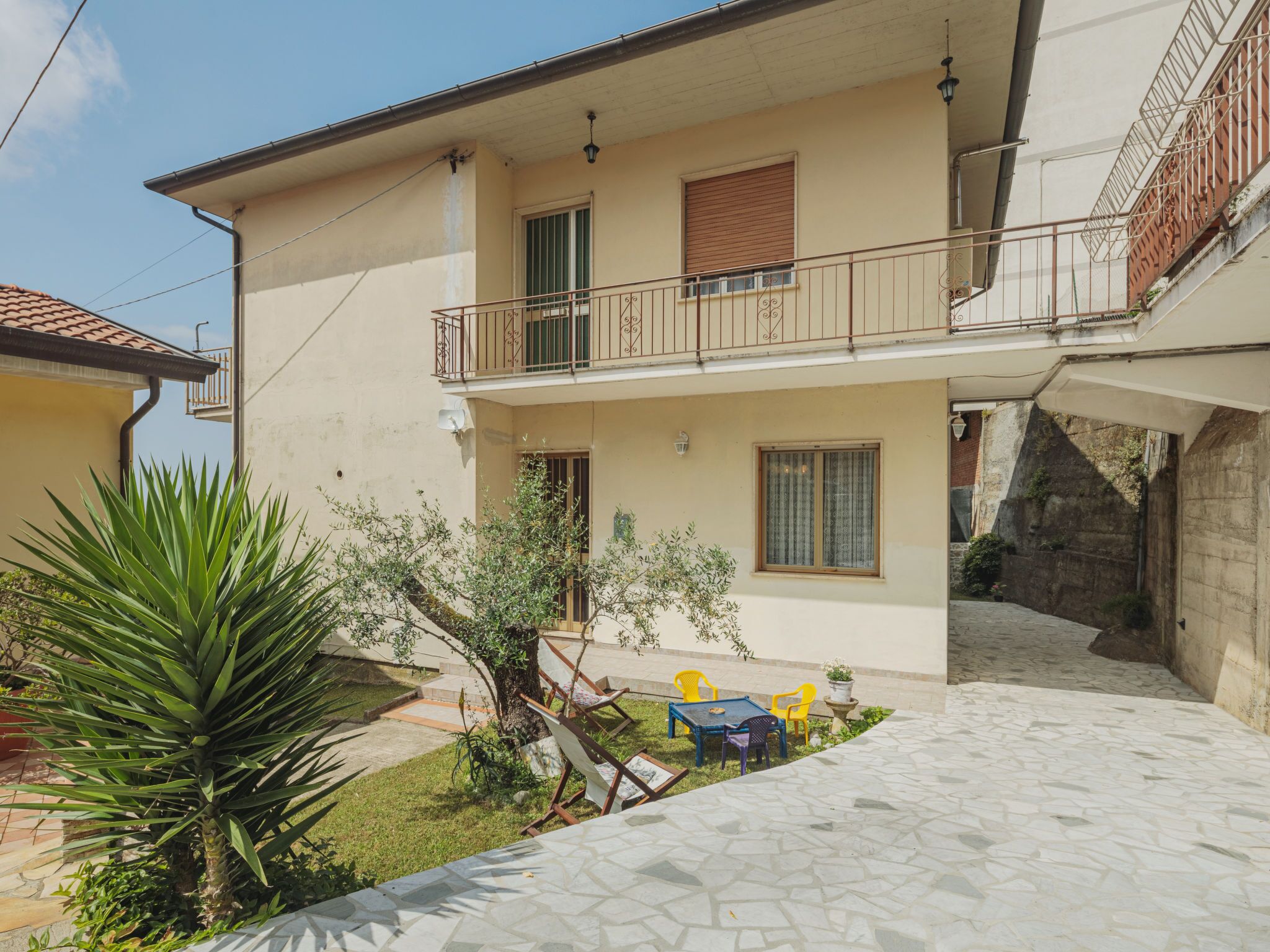 A two-story building with a balcony, a small garden with a palm tree, and a tiled patio area.