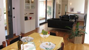Un comedor con una mesa dispuesta para dos, un armario con cristalería, y una sala de estar al fondo.