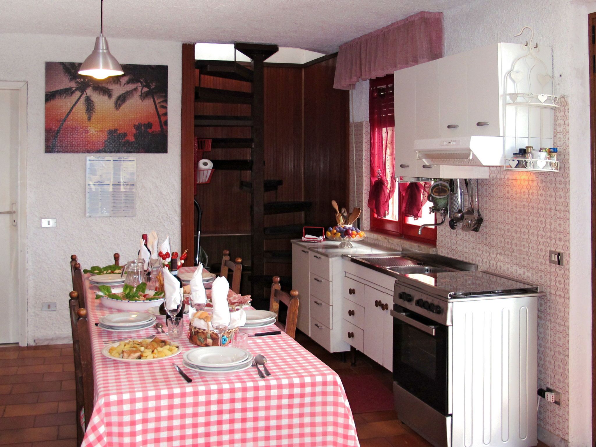 Een eethoek met een geruit tafelkleed, houten stoelen, een schilderij, en een keuken met fornuis en spoelbak.