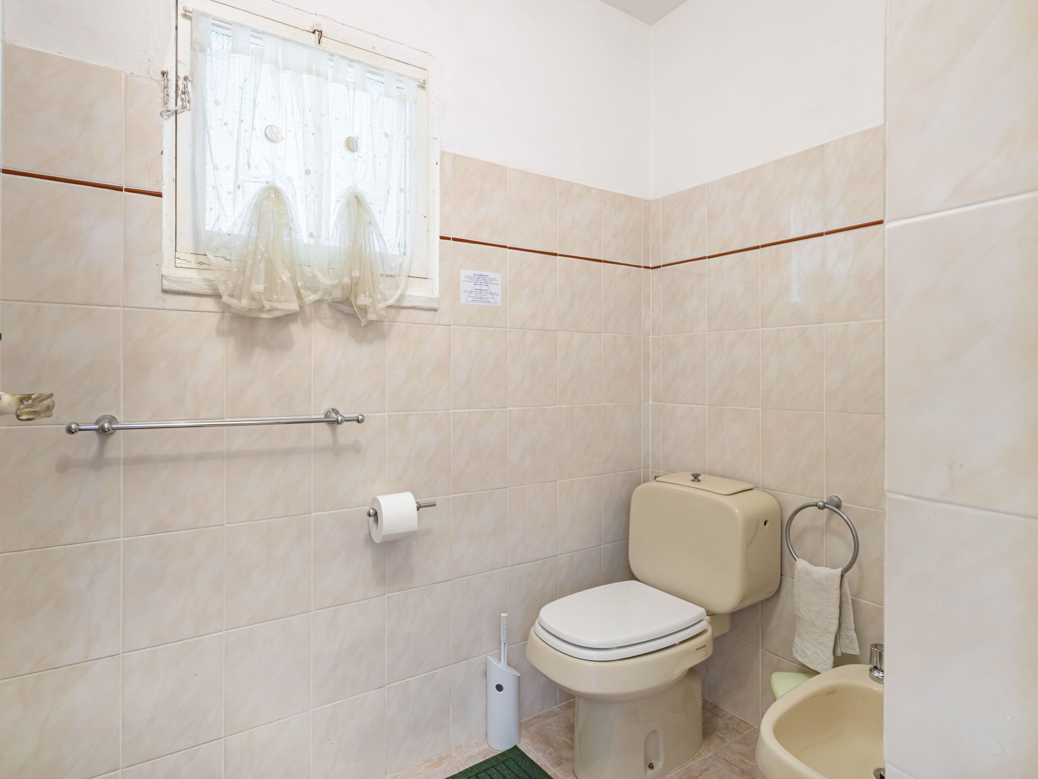 Een badkamer met beige tegels, een toilet, een bidet, een handdoekhouder en een raam met dunne gordijnen.