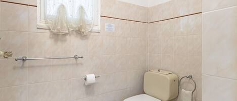 Een badkamer met beige tegels, een toilet, een bidet, een handdoekhouder en een raam met dunne gordijnen.