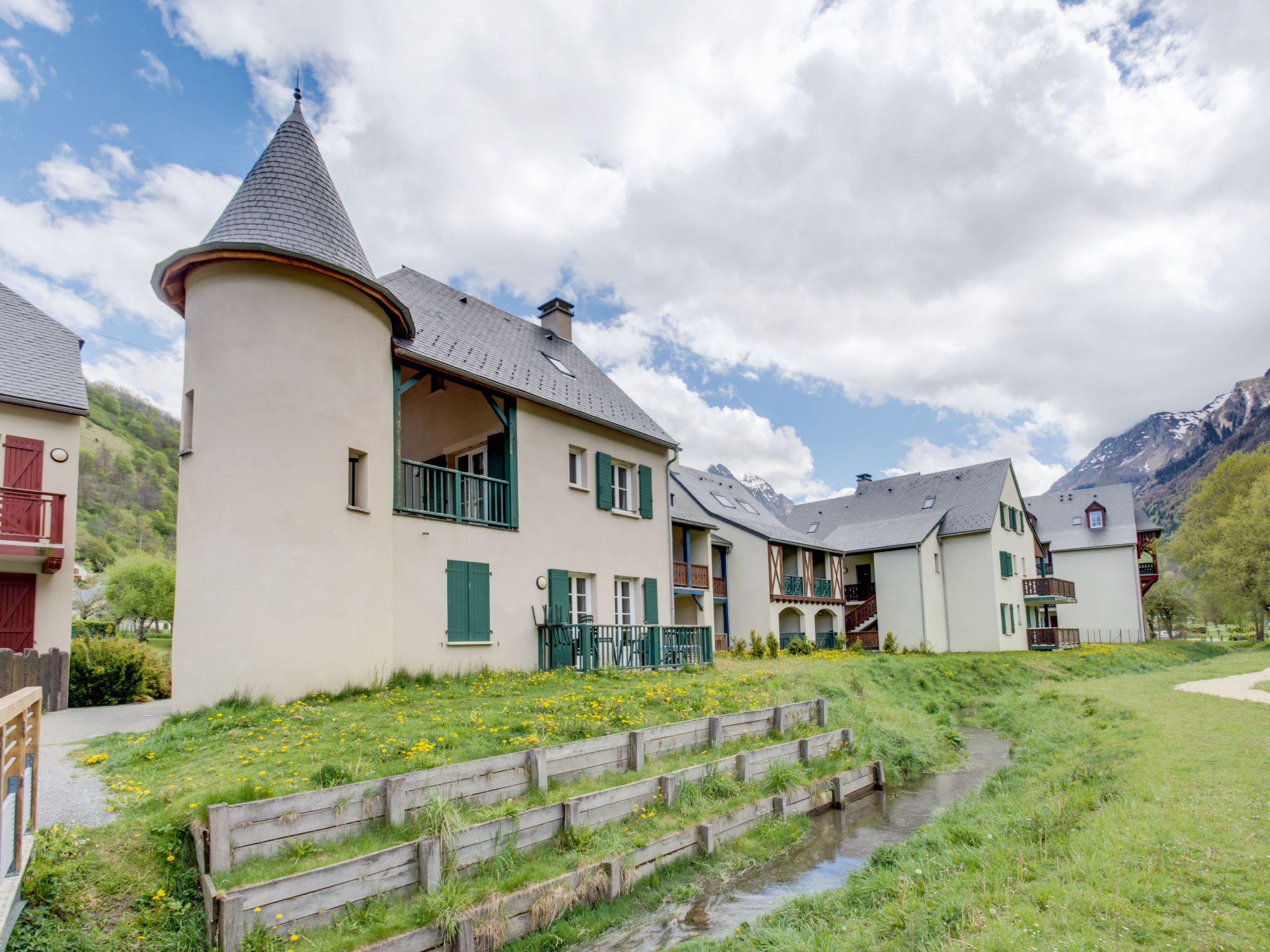 A traditional chalet-style building with a prominent tower, multiple balconies, and a small stream in the foreground.