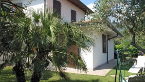 Una casa con fachada blanca, contraventanas oscuras y techo de tejas. Hay un jardín con palmeras y un columpio.