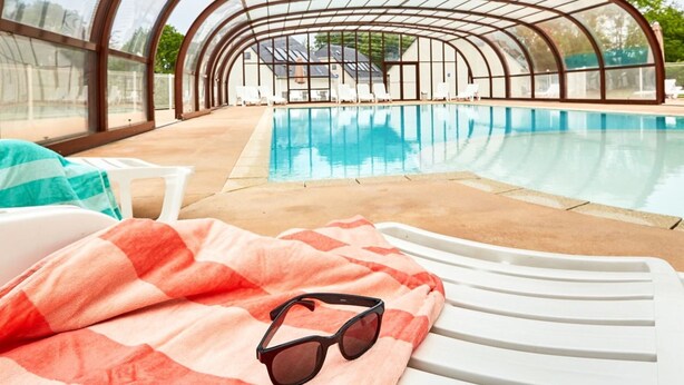 Ein Hallenbad mit Glasdach, Liegestühlen und einer Handtuch mit Sonnenbrille auf einem Stuhl.