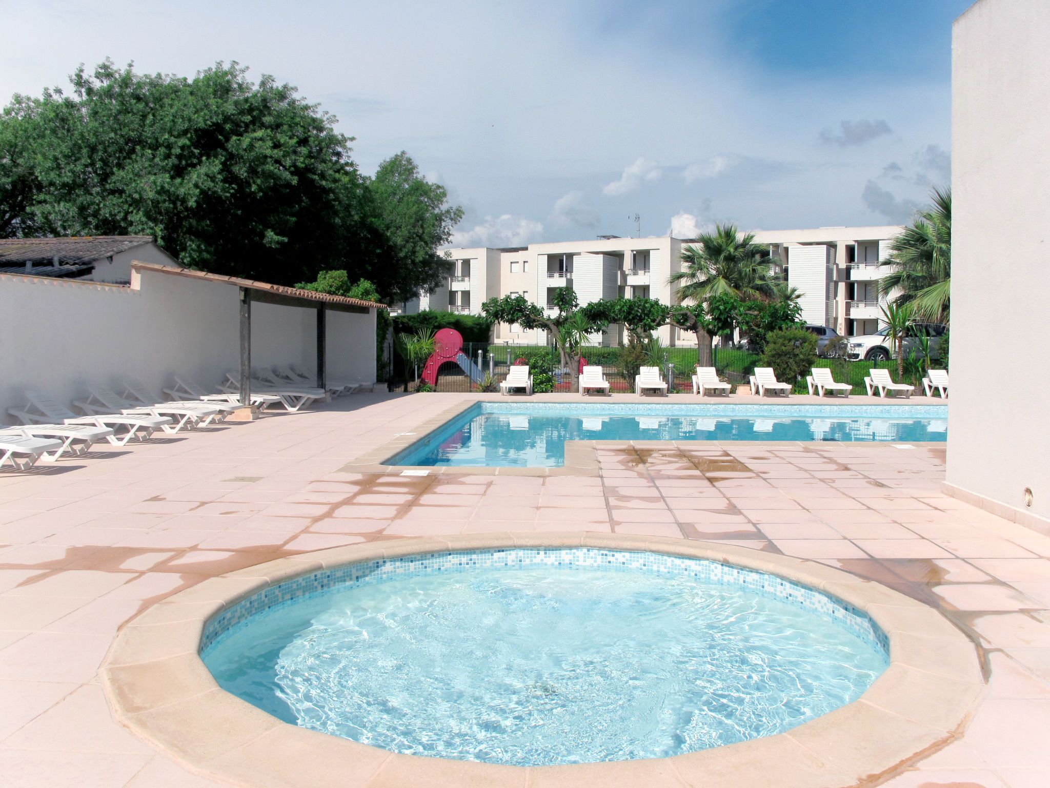 Zona d'una piscina amb gandules i un banyera d'hidromassatge, envoltada d'edificis i vegetació.