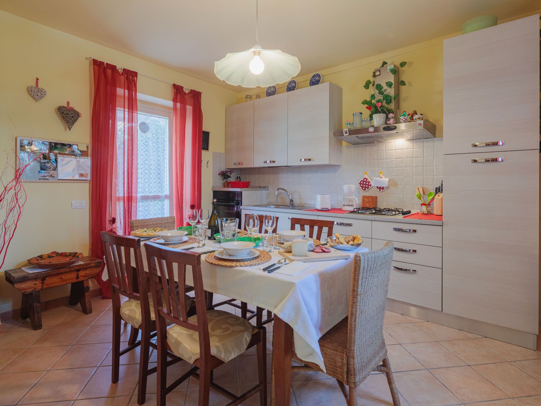 Un coin repas avec une table dressée pour quatre personnes, entourée de chaises en bois. On y trouve une cuisine avec des placards blancs et une cuisinière à gaz.