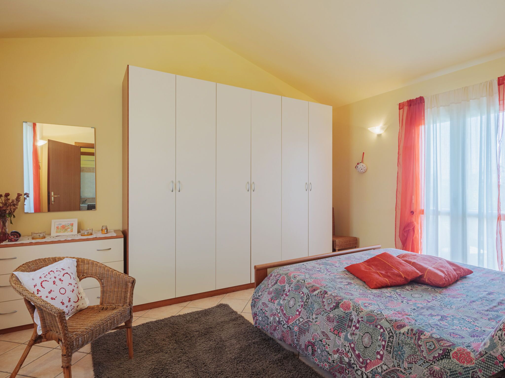 A bedroom with a bed, a wardrobe, a chair, a mirror, and a window with curtains.