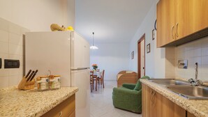 Una cocina moderna con refrigerador, lavamanos y gabinetes de madera. Se observa un comedor con mesa y sillas al fondo.