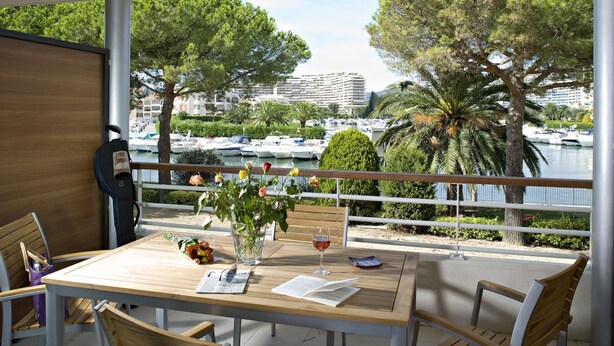 A balcony with a wooden table, chairs, a vase with flowers, and a glass of wine. A cityscape with boats and buildings is visible in the background.