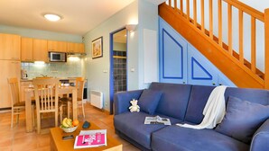 Ein Wohnzimmer mit blauem Sofa, einem Holzespeisetisch und Stühlen. Es gibt eine Küchenzeile mit Schränken und einer Mikrowelle. Im Hintergrund ist eine Treppe mit Holzbalkonen zu sehen.