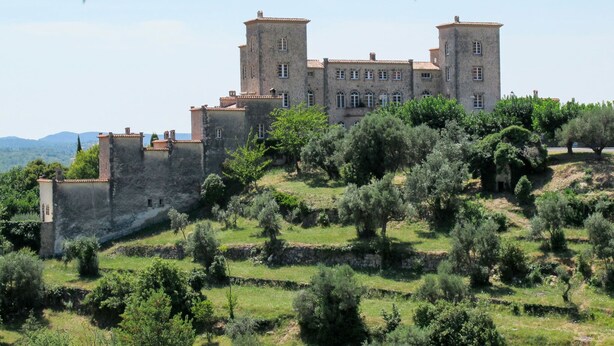 Ein historisches Steingebäude mit mehreren Türmen, umgeben von Olivenbäumen und einer hügeligen Landschaft.