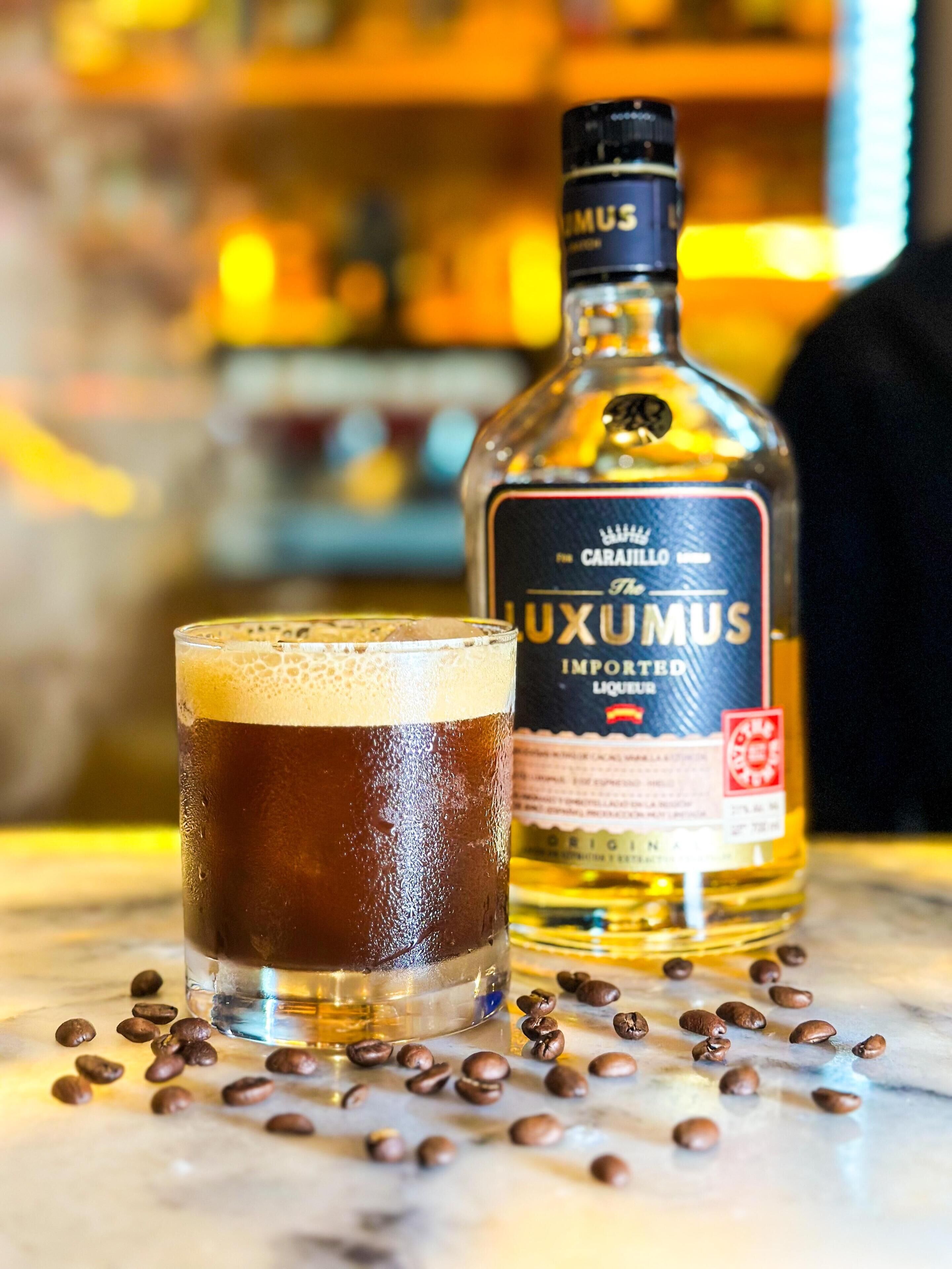Una jarra de cerveza oscura con espuma, una botella de licor importado Luximus y granos de café esparcidos sobre una superficie de mármol.