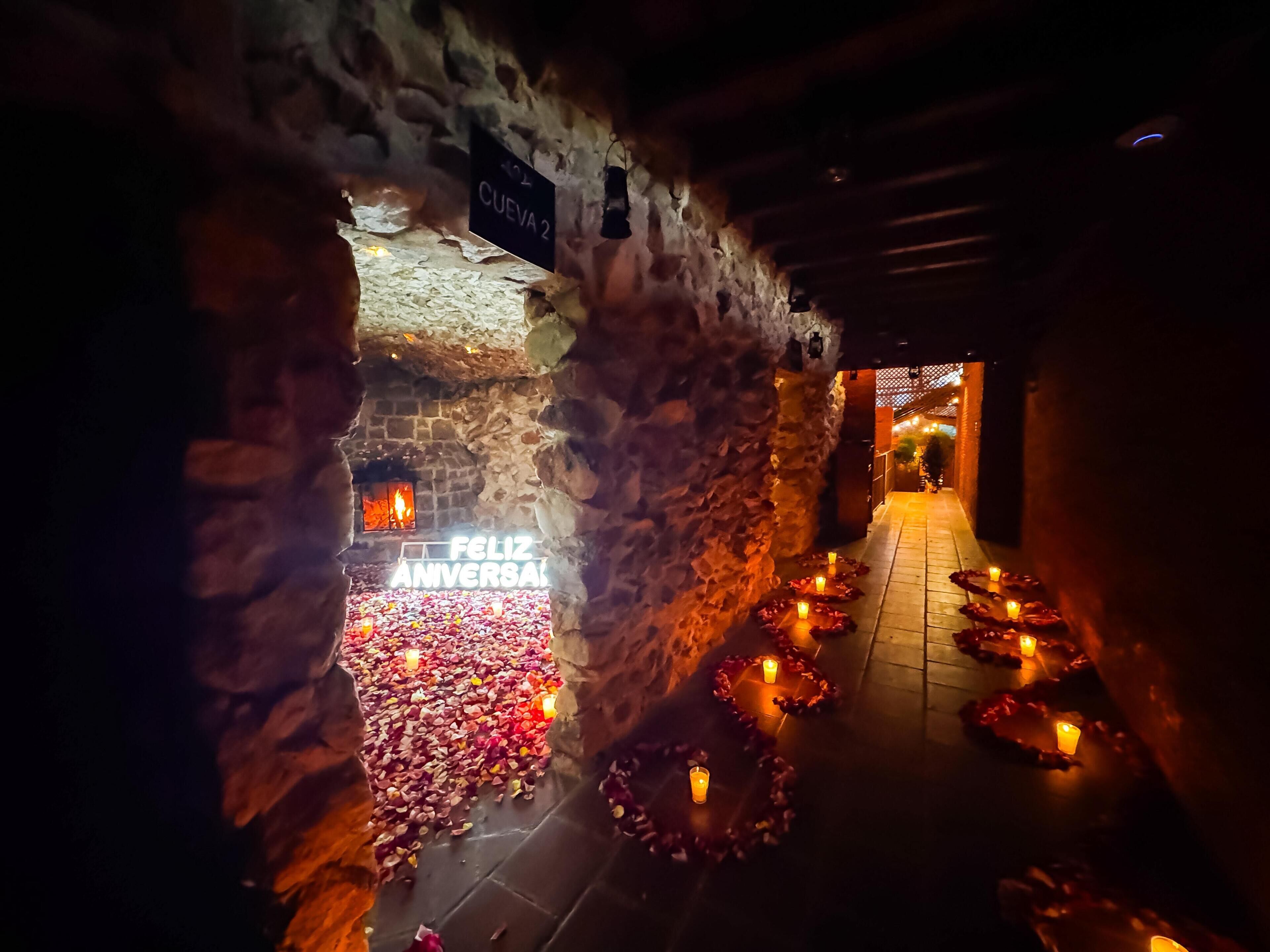A stone-walled corridor with a 'FELIZ ANIVERSARIA' sign, candles, and rose petals on the floor.