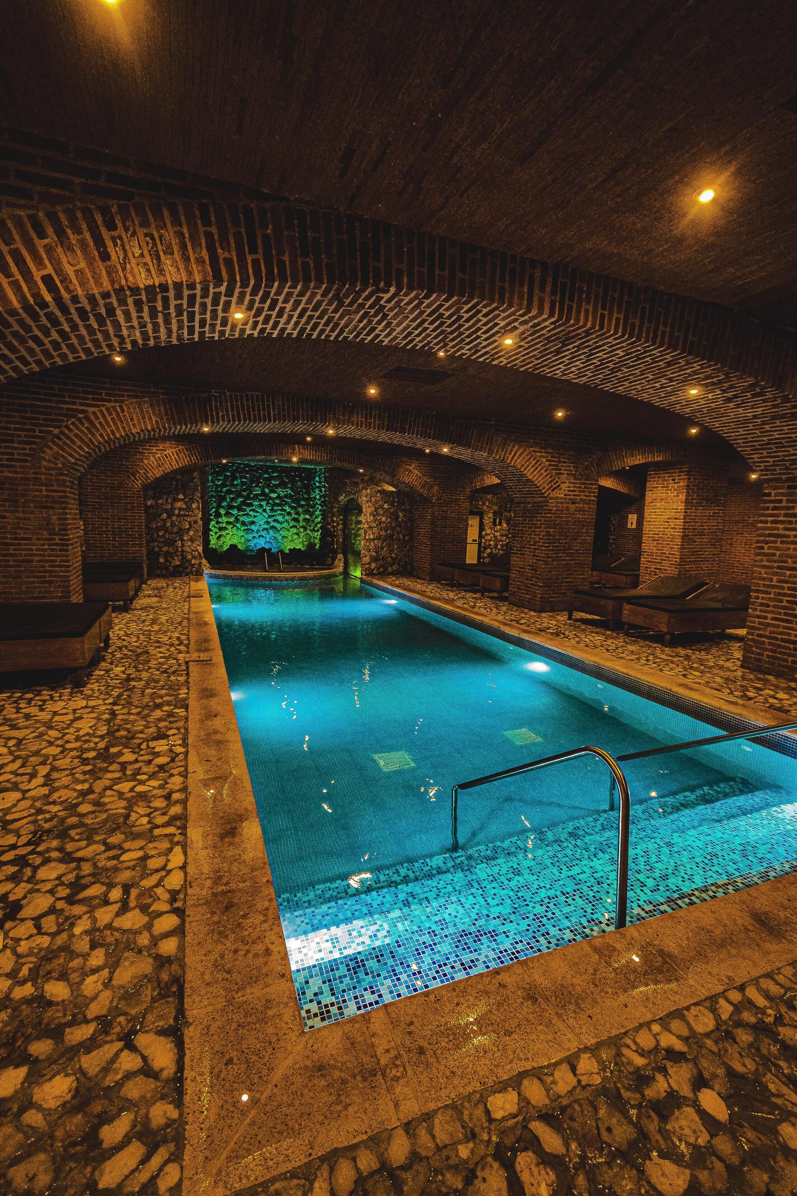 An indoor swimming pool with a stone floor and brick walls, featuring a green wall with a mosaic design at the far end.