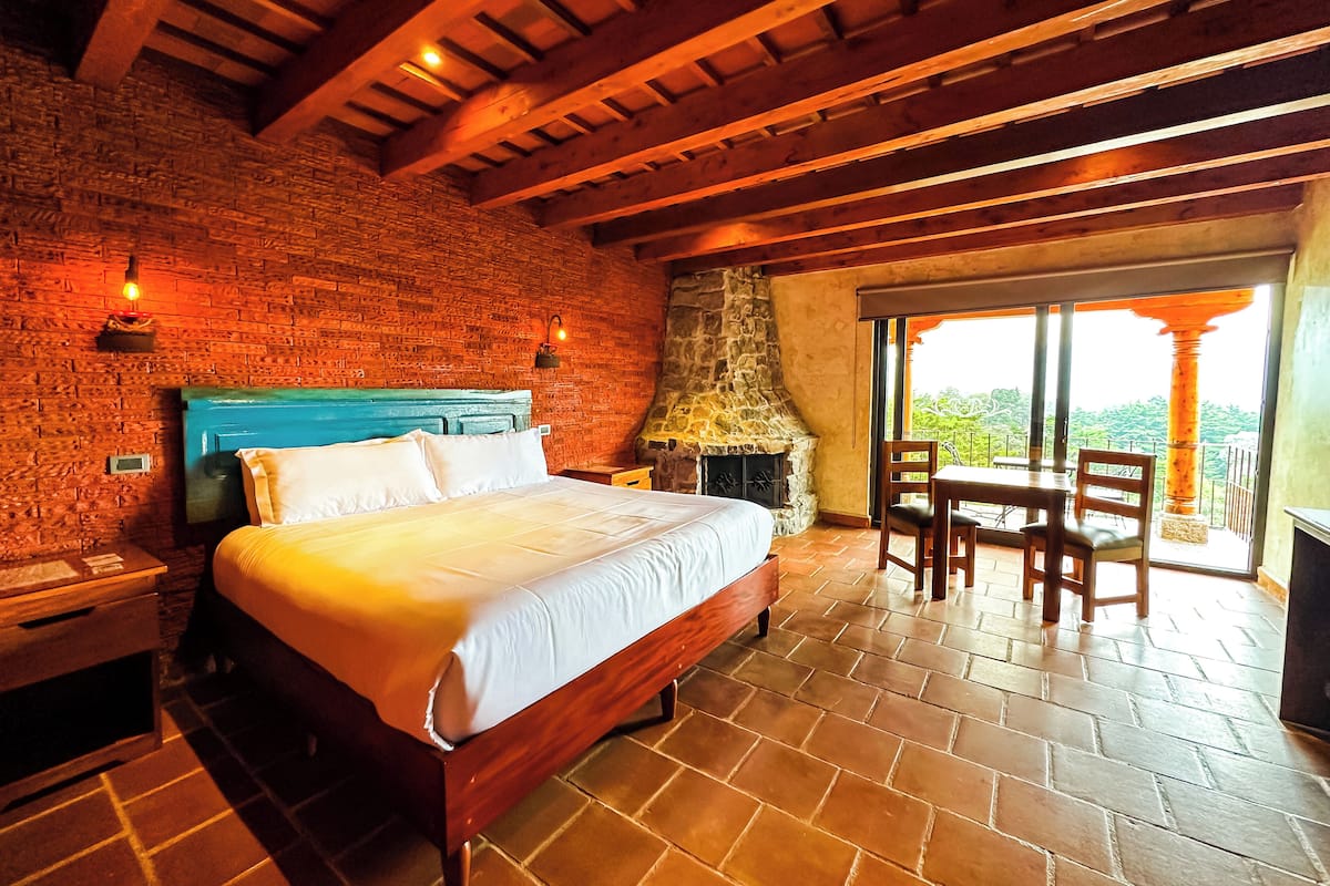 Camera da letto con parete in mattoni, soffitto in legno, camino in pietra, un letto, un tavolo con sedie e vista attraverso una porta in vetro.