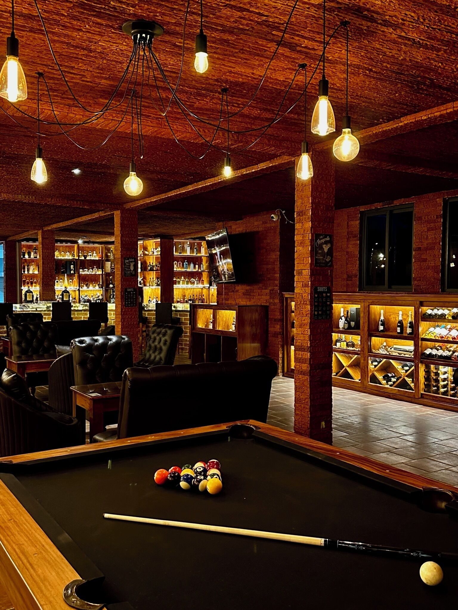 Una mesa de billar con bolas y tacos, una mesa de pool, sillones de cuero y una zona de bar con estantes llenos de botellas.