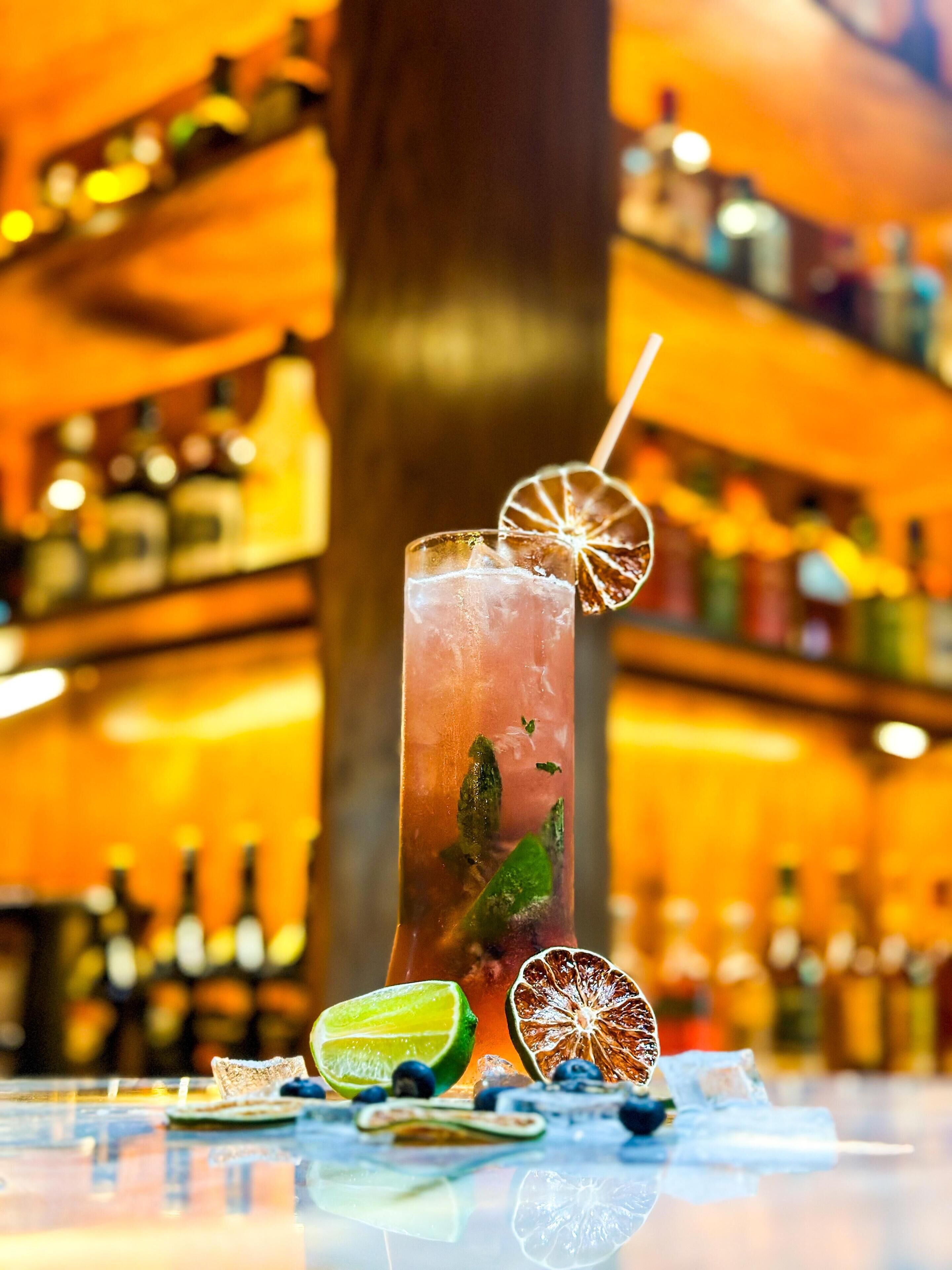 Een barsetting met een hoog glas cocktail, versierd met een plak gedroogde citrus en muntblaadjes, omringd door verschillende flessen en een partje limoen op de bar.