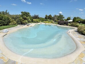 Ein klarer, ovaler Pool mit Steinrand, umgeben von üppigem Grün und blauem Himmel.