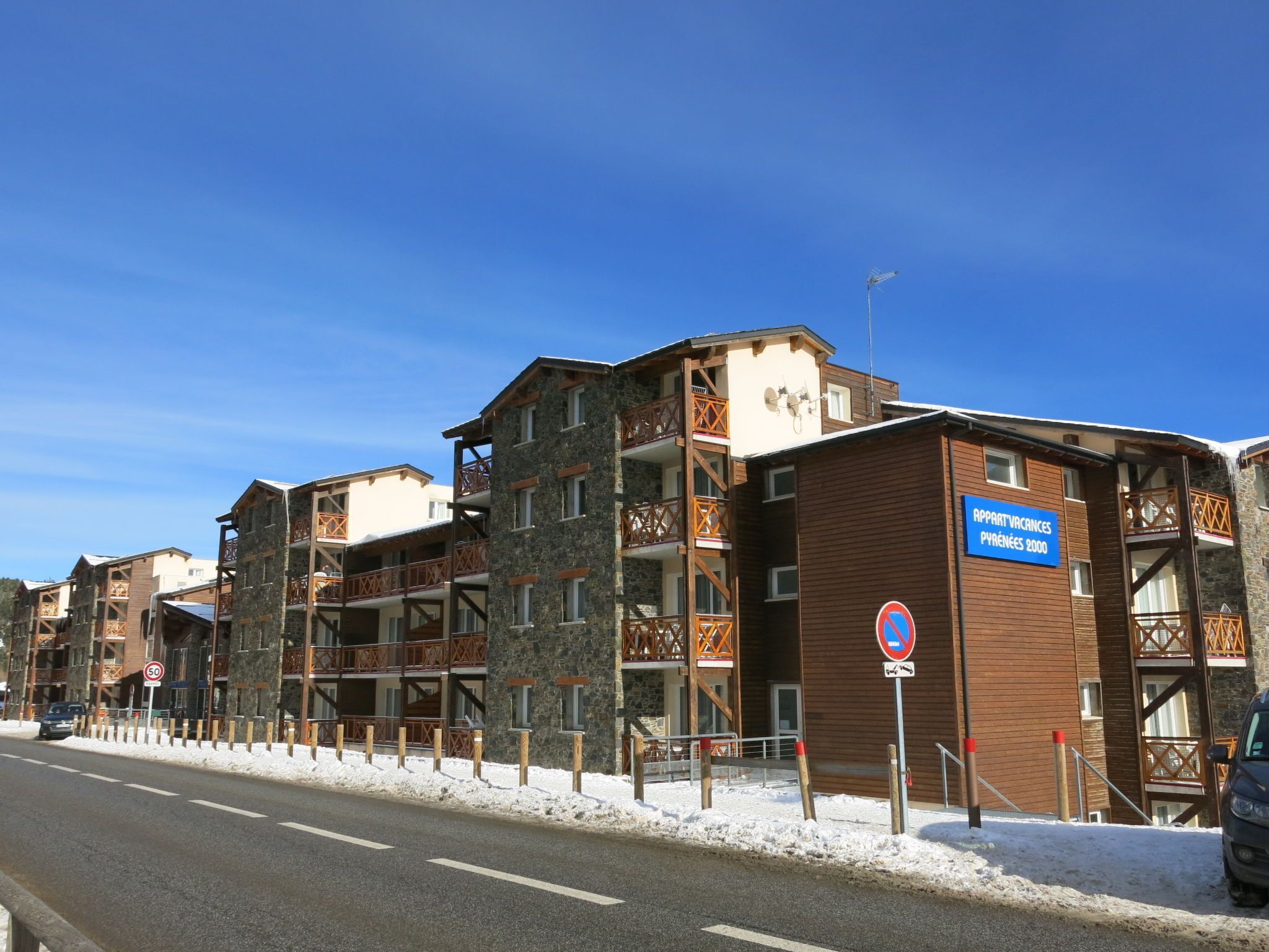 Um edifício de vários andares com fachada de pedra e varandas de madeira, uma placa indicando 'Appartments Pyrénées 2000' e uma placa de 'Proibido Estacionar' no lado.
