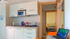 A compact kitchen with a microwave, stove, and sink, a laptop on a table, and a bed visible through an open door. - Rosolina Mare Club by Interhome (Rosolina Mare)