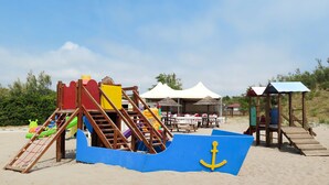 Un espace de jeux sur la plage avec un toboggan, une structure d’escalade et un tapis de jeu bleu en forme de bateau, orné d’une ancre.