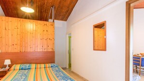 Une chambre avec un plafond en bois, un lit recouvert d’une couette à rayures colorées, une table de chevet avec une lampe et un miroir encadré en bois accroché au mur.