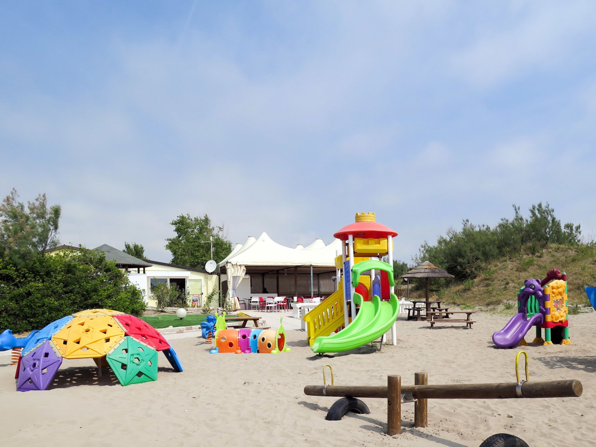 Ein Strand mit bunten Spielgeräten, darunter eine Rutsche und Klettergerüste, sowie ein Zeltenbereich im Hintergrund.