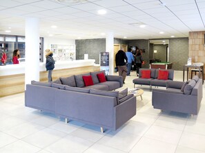 A modern lobby with grey sofas and red cushions, a pool table, and a reception area with a counter and shelves.