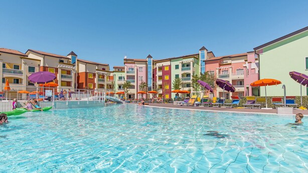 A swimming pool area with colorful umbrellas and lounge chairs, surrounded by multi-story buildings with balconies.