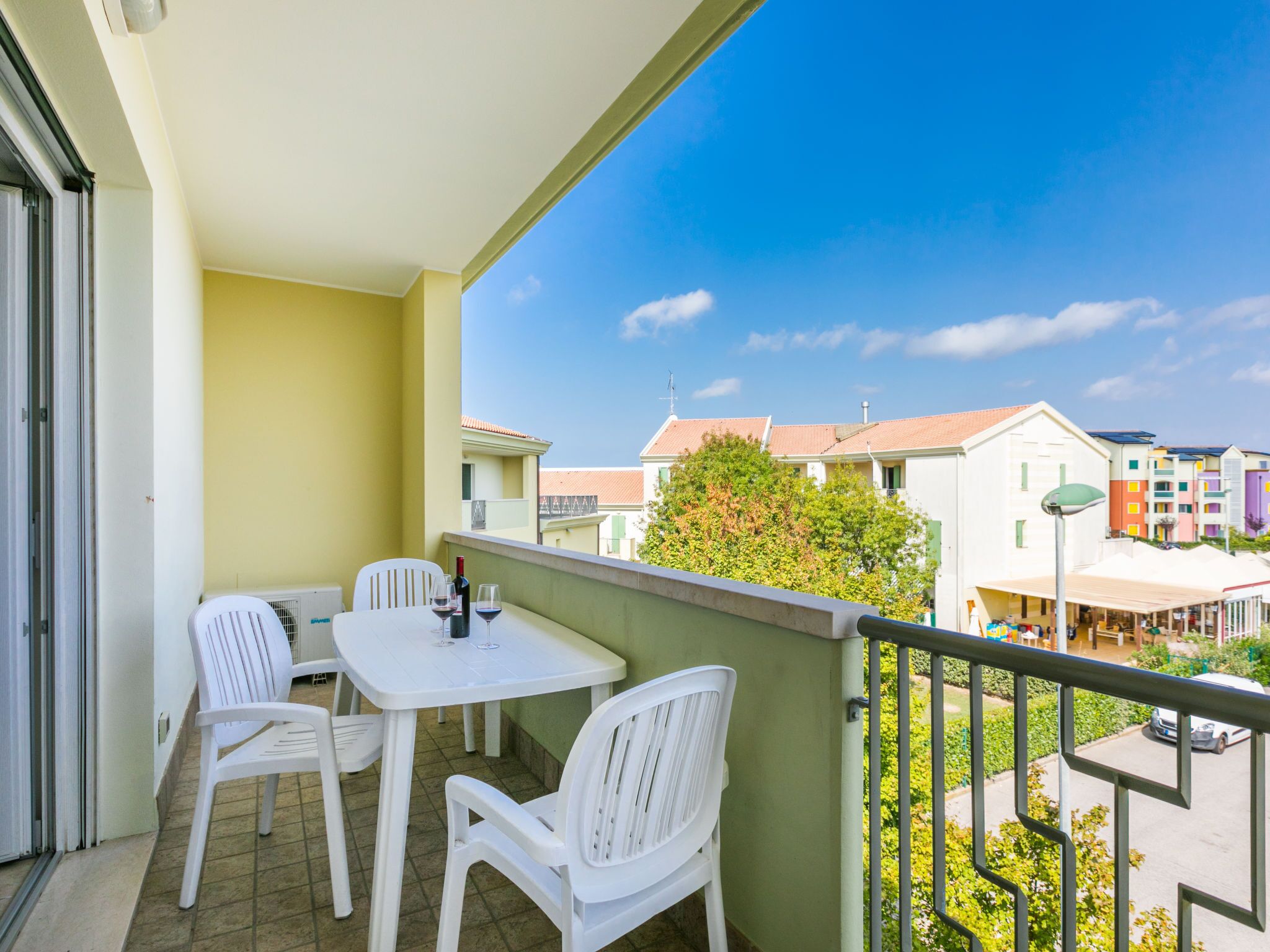 Ein Balkon mit einem weißen Tisch und zwei weißen Stühlen, mit Blick auf ein Wohngebiet mit bunten Gebäuden und blauem Himmel.