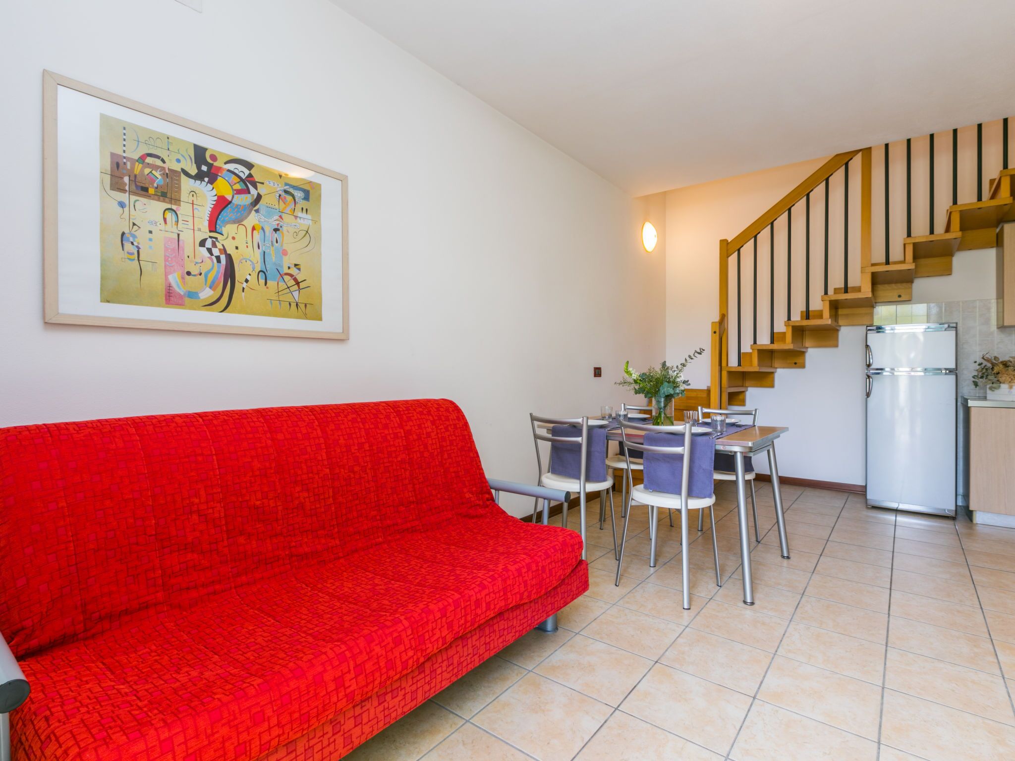 A living room with a red sofa, a dining table with chairs, a kitchen area with a refrigerator, and a staircase.