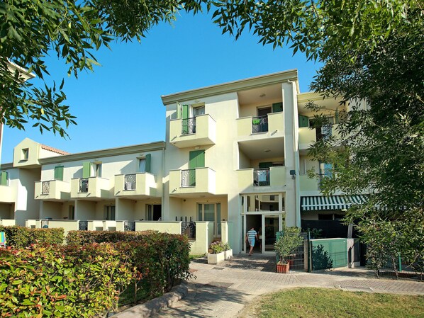 Un edificio residencial de varios pisos con balcones, entrada pavimentada y entorno con vegetación.