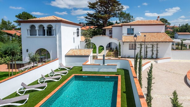 Una piscina con solárium de baldosas, rodeada de edificios blancos con ventanas de arco y techo de tejas.