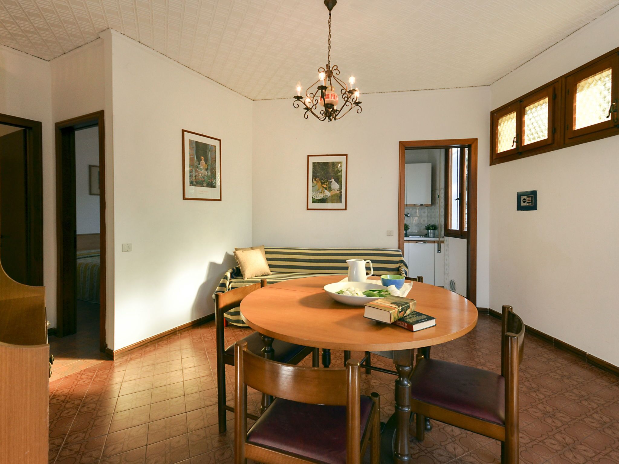 A dining area with a round wooden table, chairs, a chandelier, and a kitchen in the background.