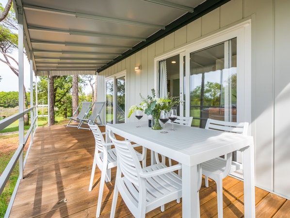 Un patio techado al aire libre con una mesa y sillas blancas, una terraza de madera y vistas a la vegetación circundante.