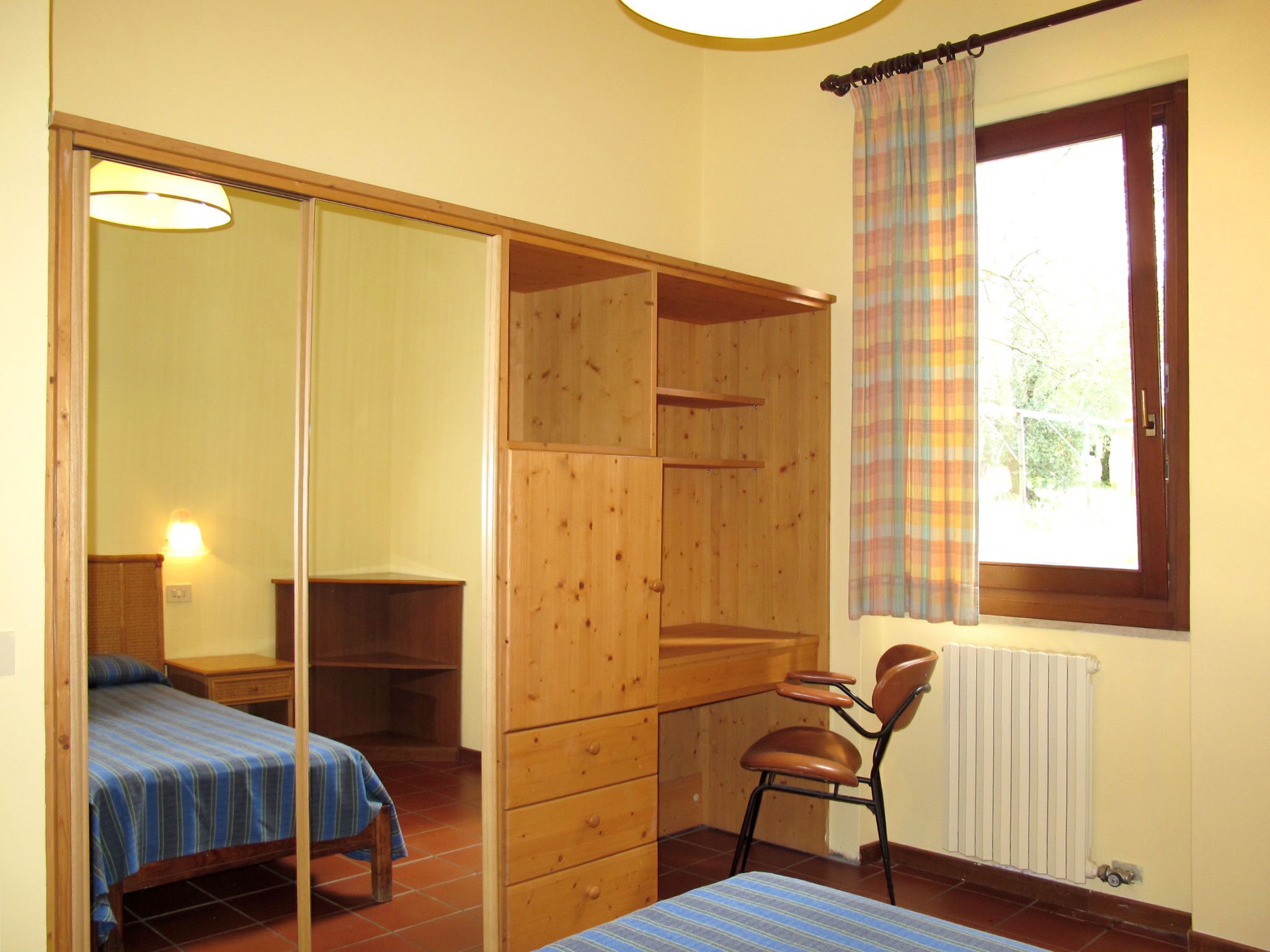 Una habitación con cama, una silla, armario de madera, radiador y ventana con cortinas.
