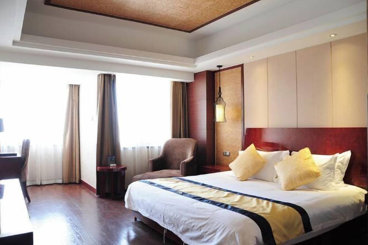 Quarto de hotel com uma cama grande, uma escrivaninha e uma cadeira. Há uma janela com cortinas, uma luminária na parede e uma cabeceira de madeira.