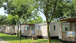 Une rangée de maisons mobiles dotées de terrasses en bois et de volets verts, entourées d’arbres et bordées d’un chemin pavé.