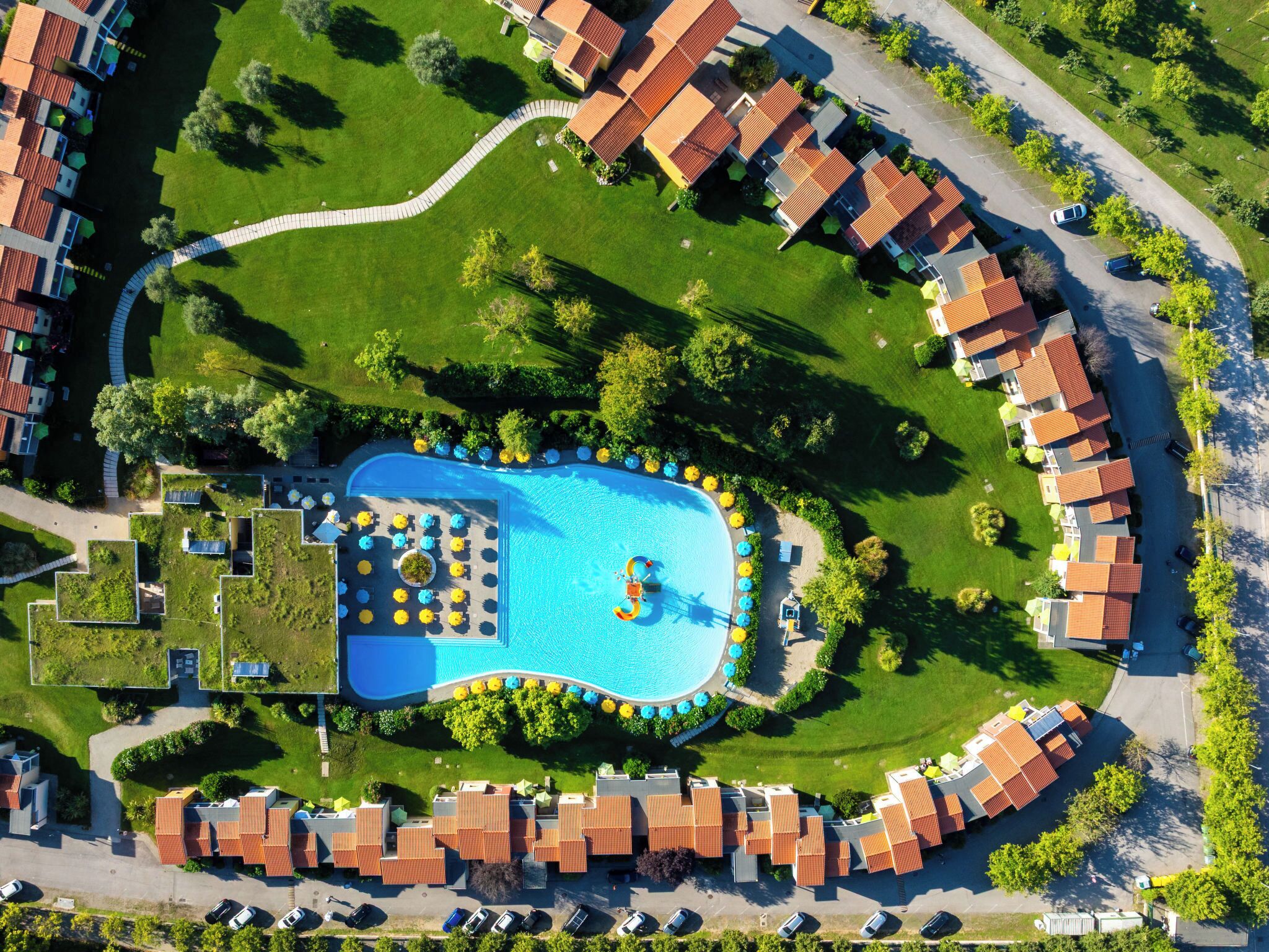 An aerial view of a residential area with a swimming pool, green lawns, and uniform red-roofed buildings.