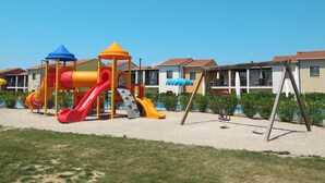 Ein Spielplatz mit Rutschen und Klettergerüsten, umgeben von Häusern mit Balkonen und Sonnenschirmen.