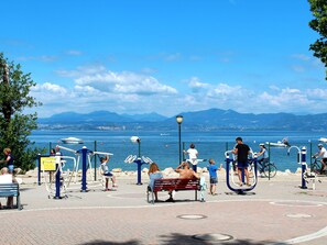 Um parque público com equipamentos de exercícios, bancos e vista para um lago com barcos.