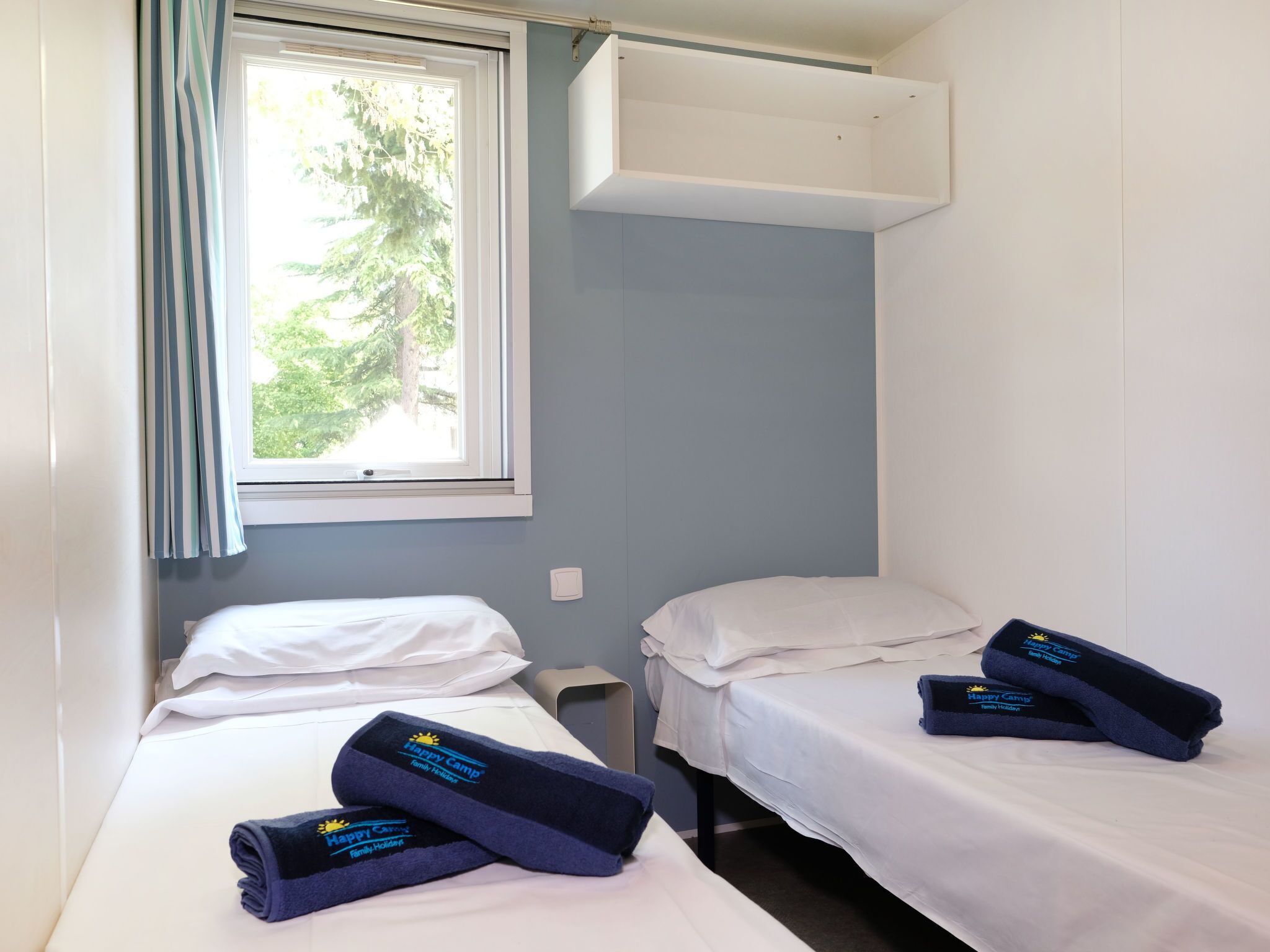 A small, single-room accommodation with two beds, a window with curtains, and a white wardrobe.