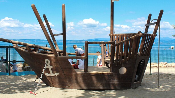 Eine hölzerne Piratenschiff-Konstruktion mit Flagge, auf einem Strand mit freiem Blick auf das Meer und ferne Berge.