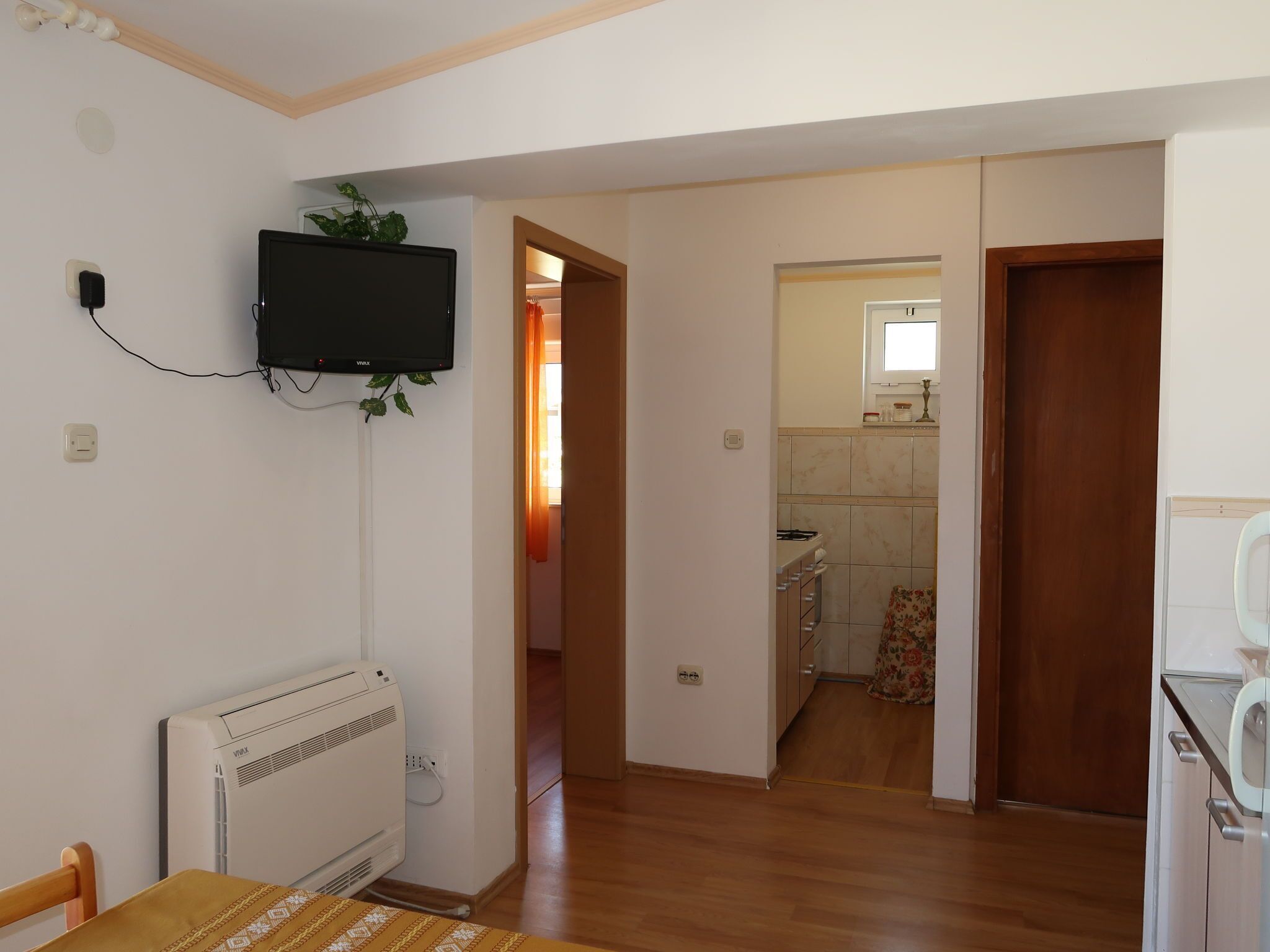 Een kamer met een aan de muur bevestigde televisie, een radiator, een houten tafel en een keukenruimte zichtbaar door een geopende deur.