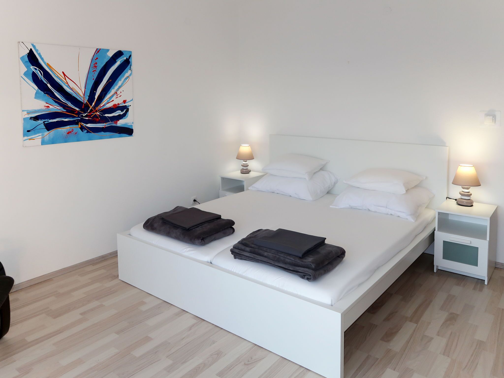 Un dormitorio moderno con una cama blanca, dos mesitas de noche con lámparas y un cuadro abstracto grande en la pared.