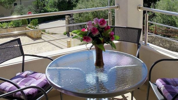 Uma mesa de vidro redonda com um vaso de flores, rodeada por duas cadeiras com almofadas roxas, numa varanda com vista para a costa.