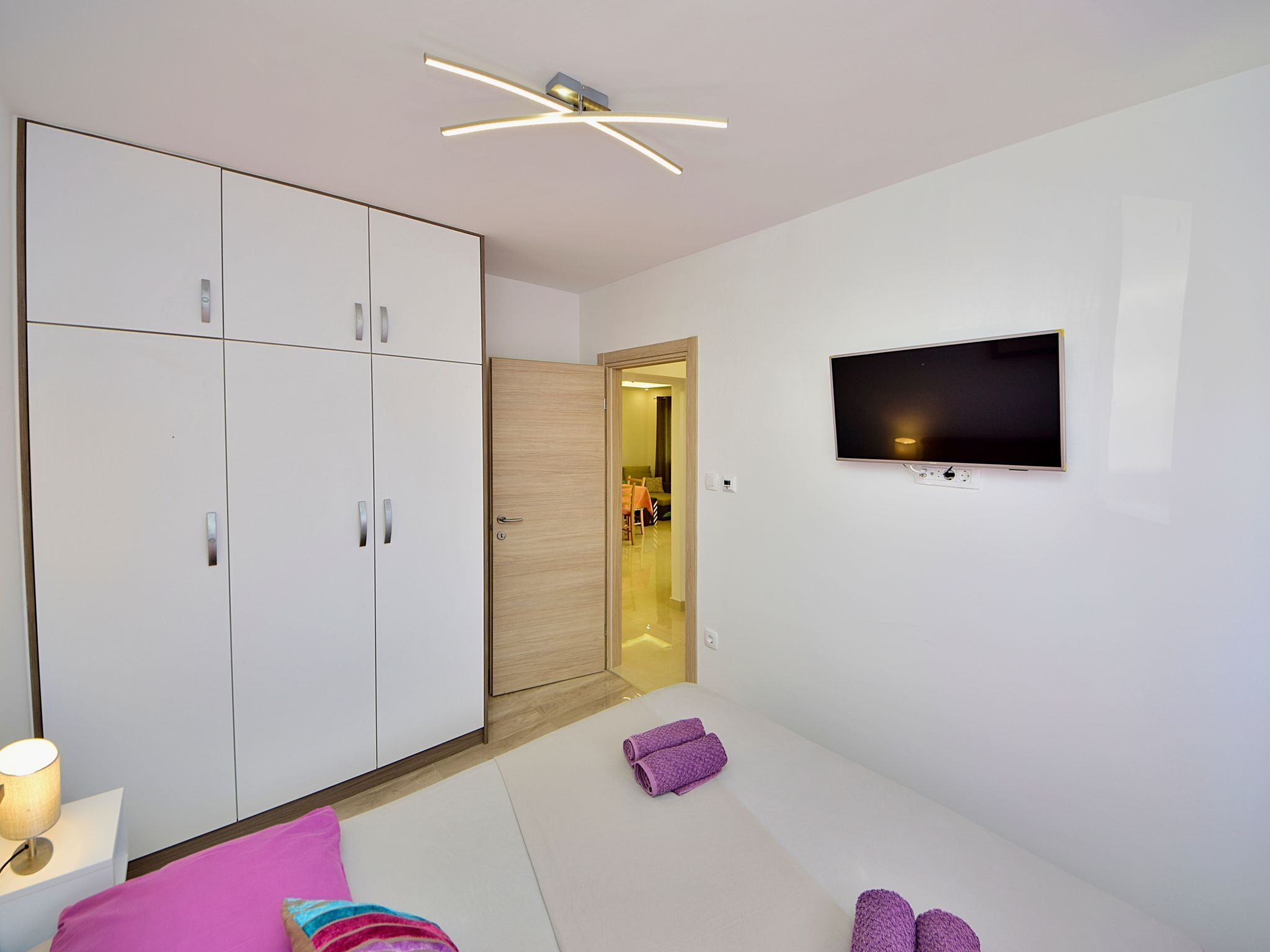 Ein modernes Hotelzimmer mit großem Kleiderschrank, einem flachbildfernseher an der Wand und einem Bett mit einer Decke in Rosa und Blau.
